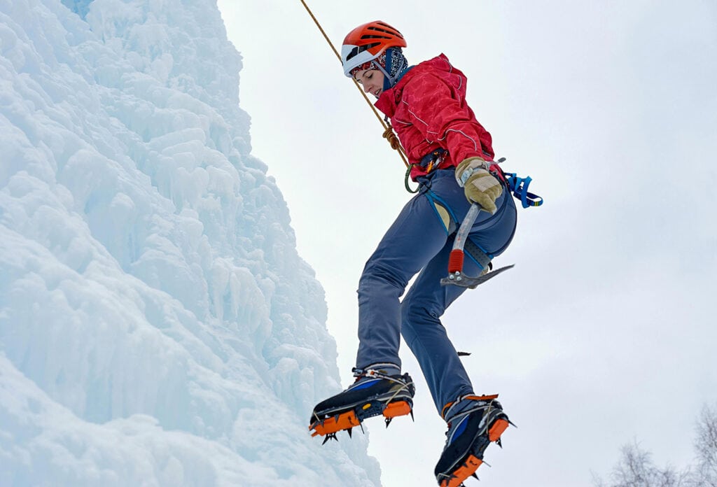 Alpinist woman with ice axe and orange helmet climbing a large ice wall