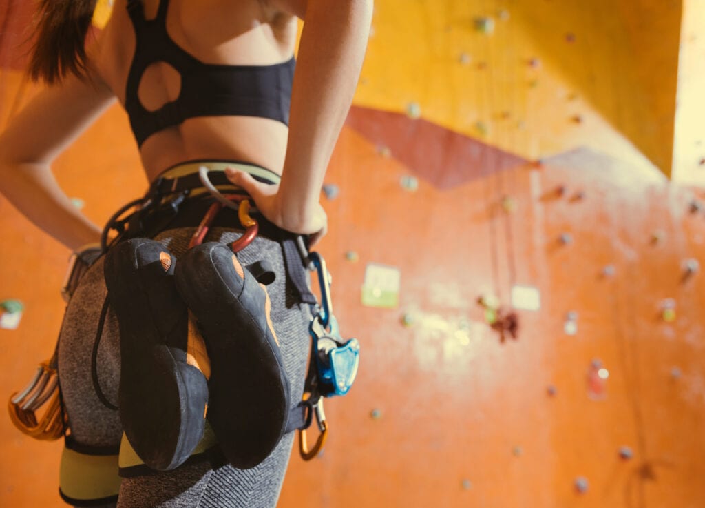 woman indoor rock climbing with a pair of climbing shoes clipped to her harness