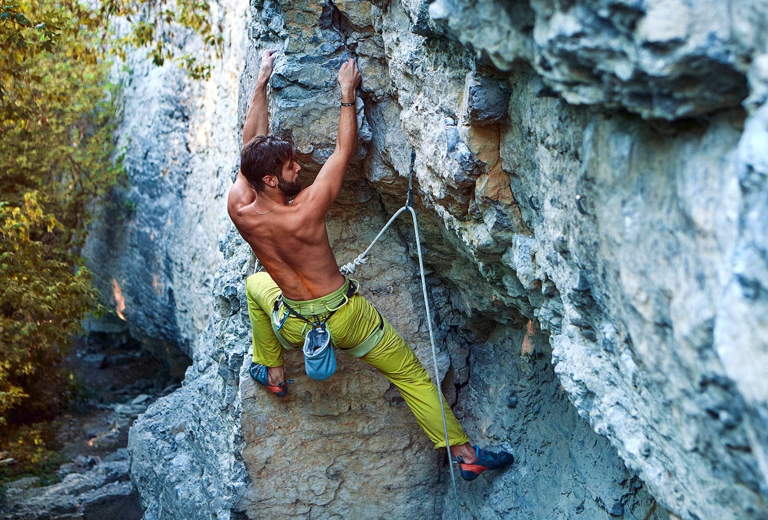 muscular man rock climber in bright yellow pants climbing the challenging route on the cliff in forest